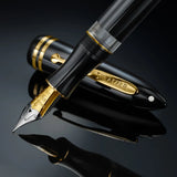 Black fountain pen with gold trim and a steel nib, the cap engraved SHEAFFER, resting on a glossy black reflective surface